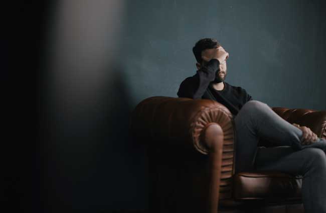 image of depressed man in sweatpants sitting on a couch in a dark room with his forhead in his hand for blog post for Gulf Breeze Recovery non-12 step treatment holistic drug and alcohol rehab
