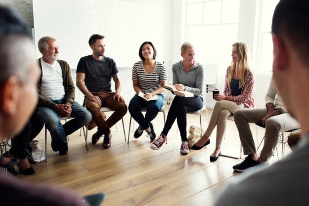 large group of men and women sitting in a circle in a well lit room while a therapist leads them in the benefits of group counseling
