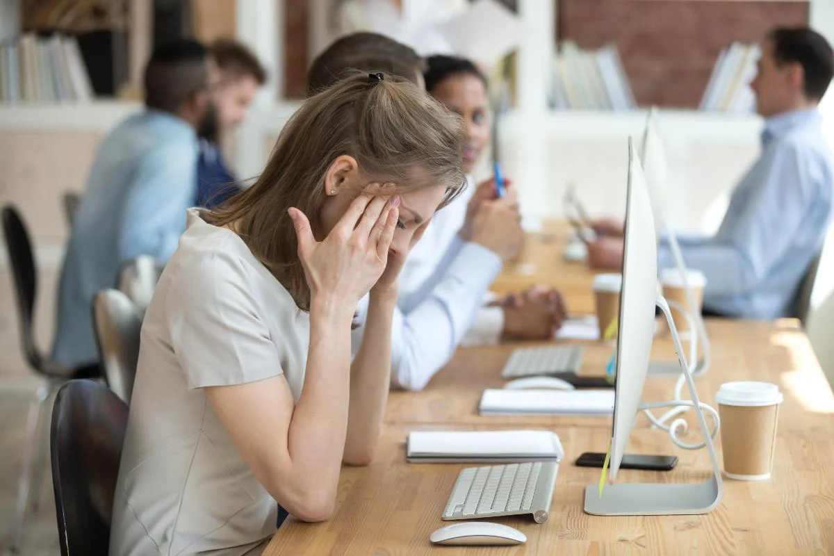 young woman in open office setting staring at computer screen while holding her head and recognizing addiction in the workplace