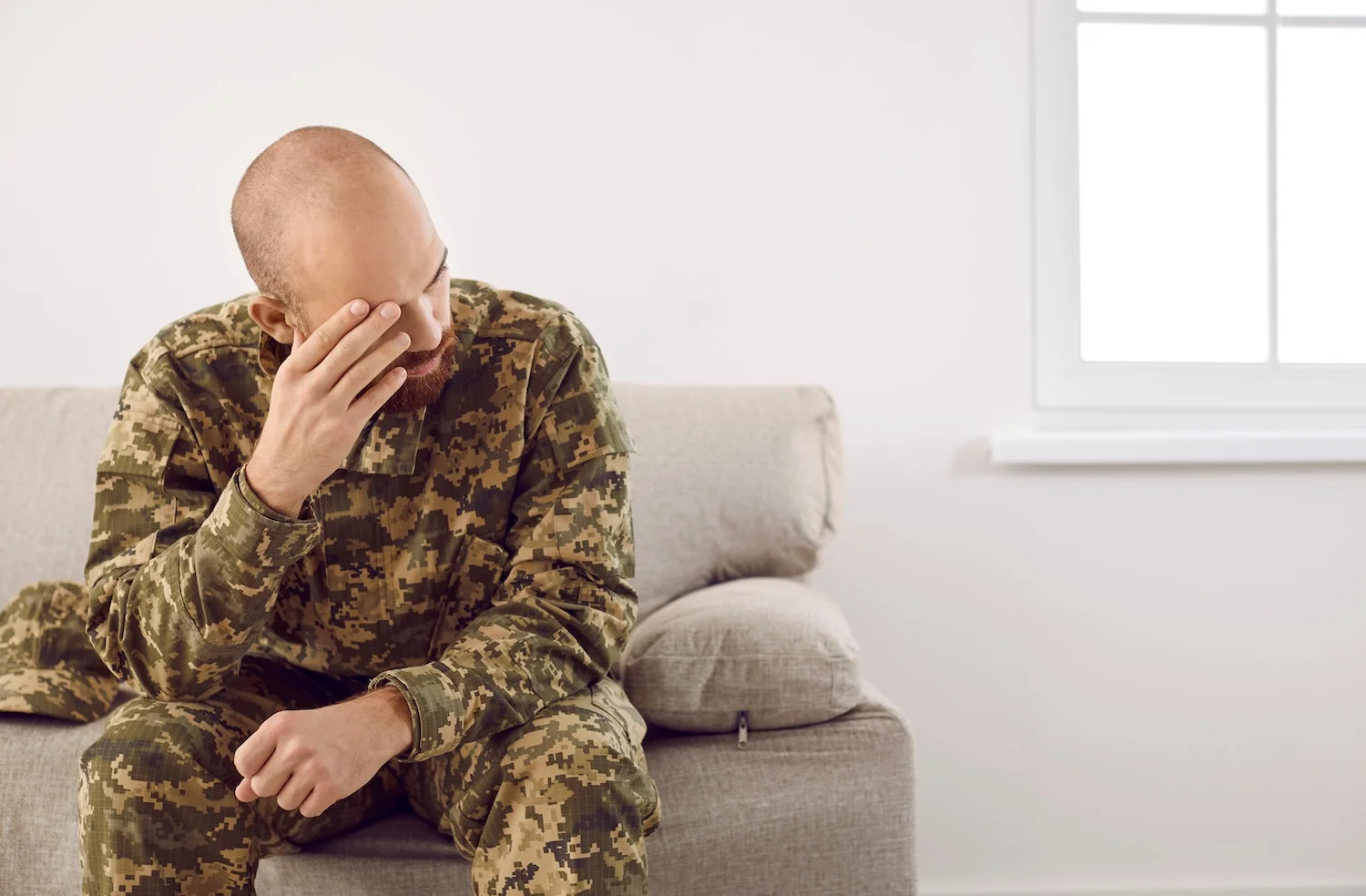 Soldier in camouflage uniform sitting on a couch indoors, leaning forward with a hand on his forehead, looking distressed or deep in thought.