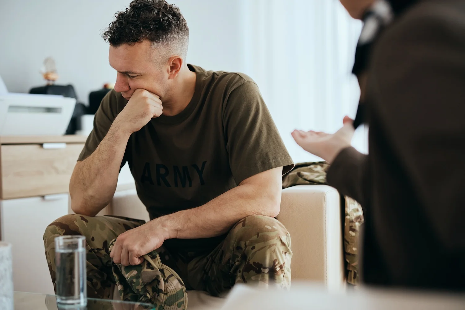 Soldier in camouflage pants and an "ARMY" t-shirt sitting on a chair, leaning forward with a hand on his chin, appearing troubled while talking to another person during a counseling session