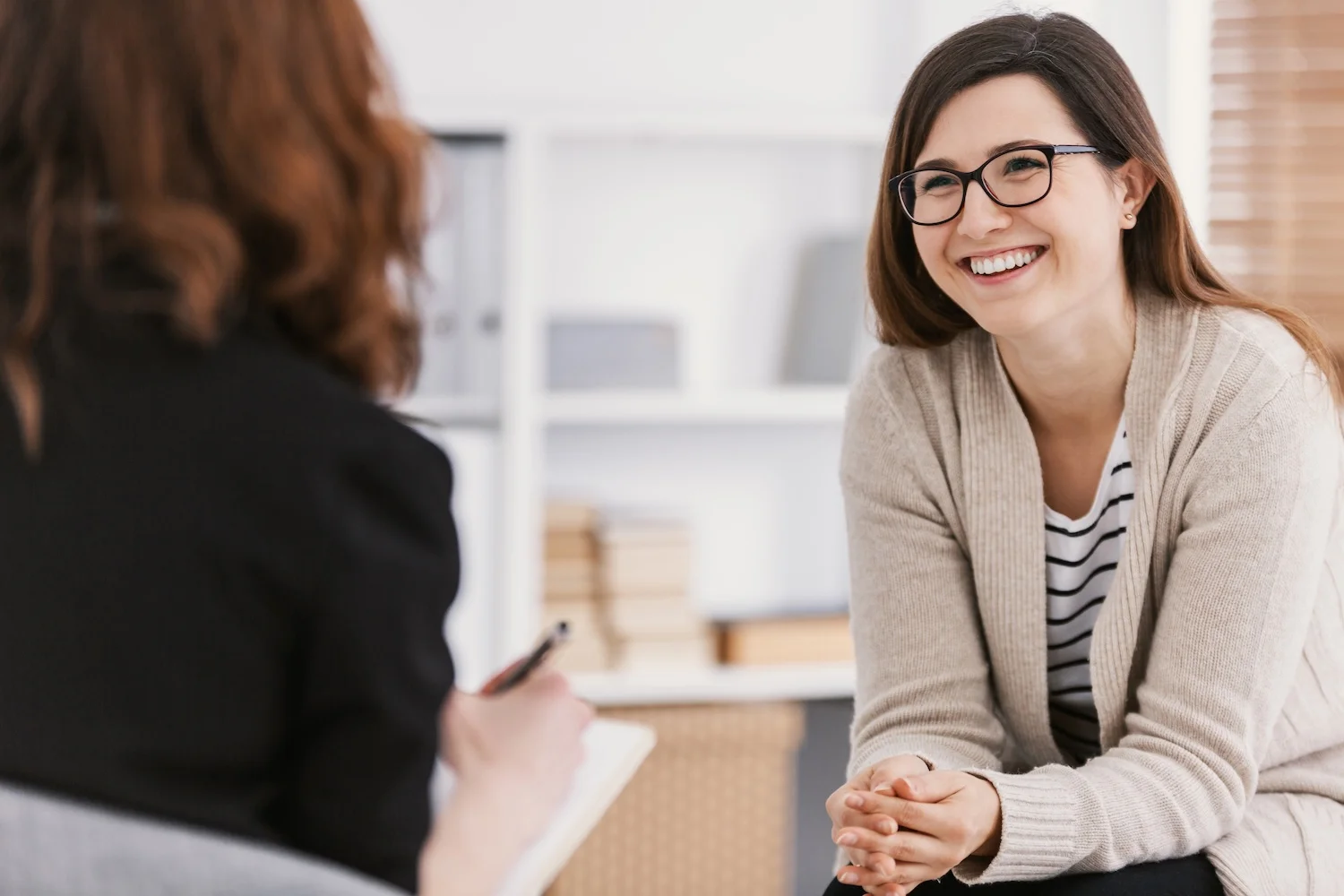 A woman sits on a chair and smiles warmly while talking with her therapist who is taking notes.